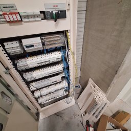 Damian Ławniczek-Wertepny - Otwarta szafa elektryczna z widocznymi zabezpieczeniami, licznikiem i okablowaniem. Na podłodze narzędzia i elementy obudowy. Widok z góry, oświetlenie sufitowe.