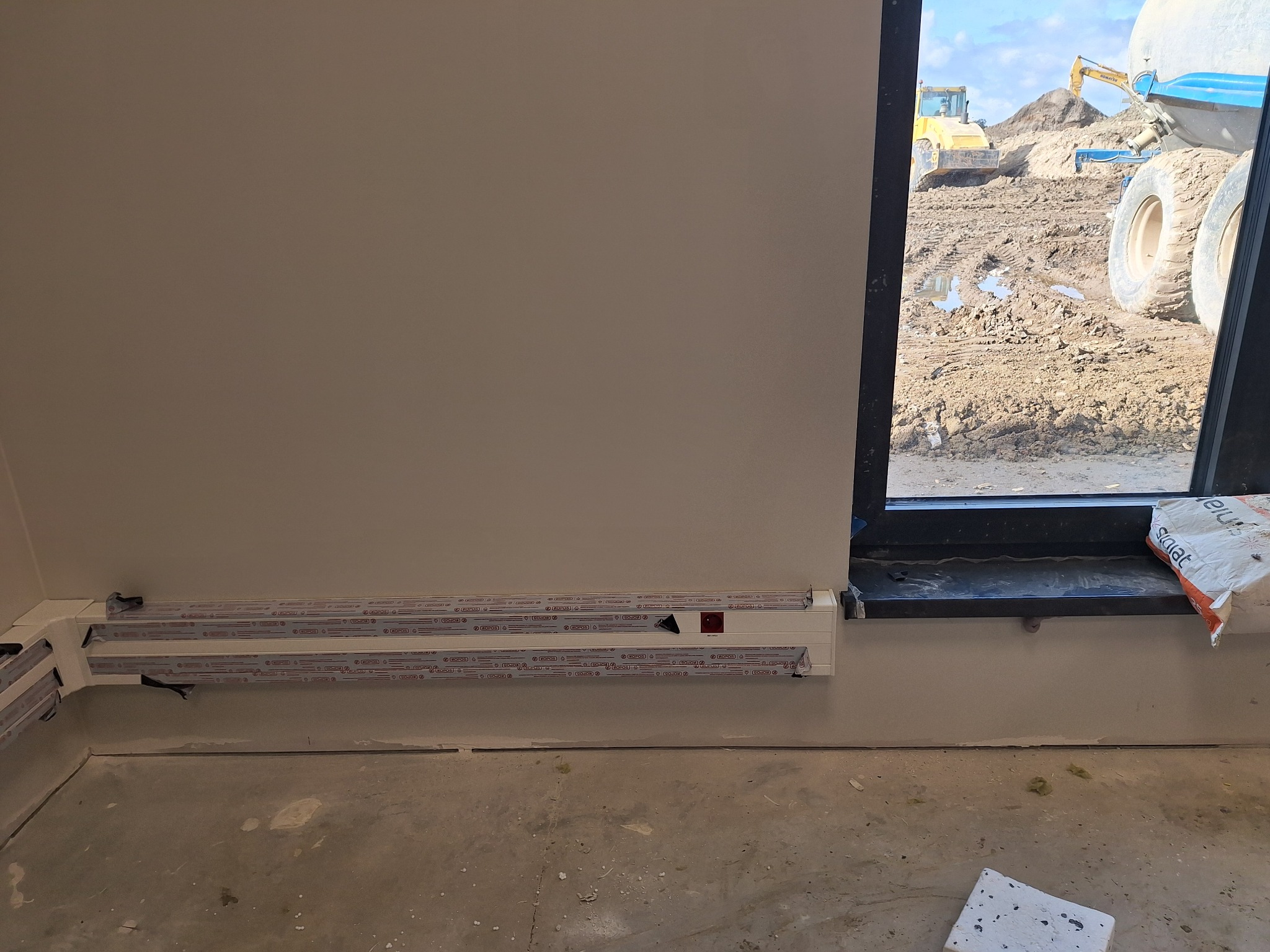 Przyścienne koryto instalacyjne z puszkami pod oknem w surowym wnętrzu. Za oknem plac budowy z maszynami. Widoczne elementy instalacji elektrycznej.