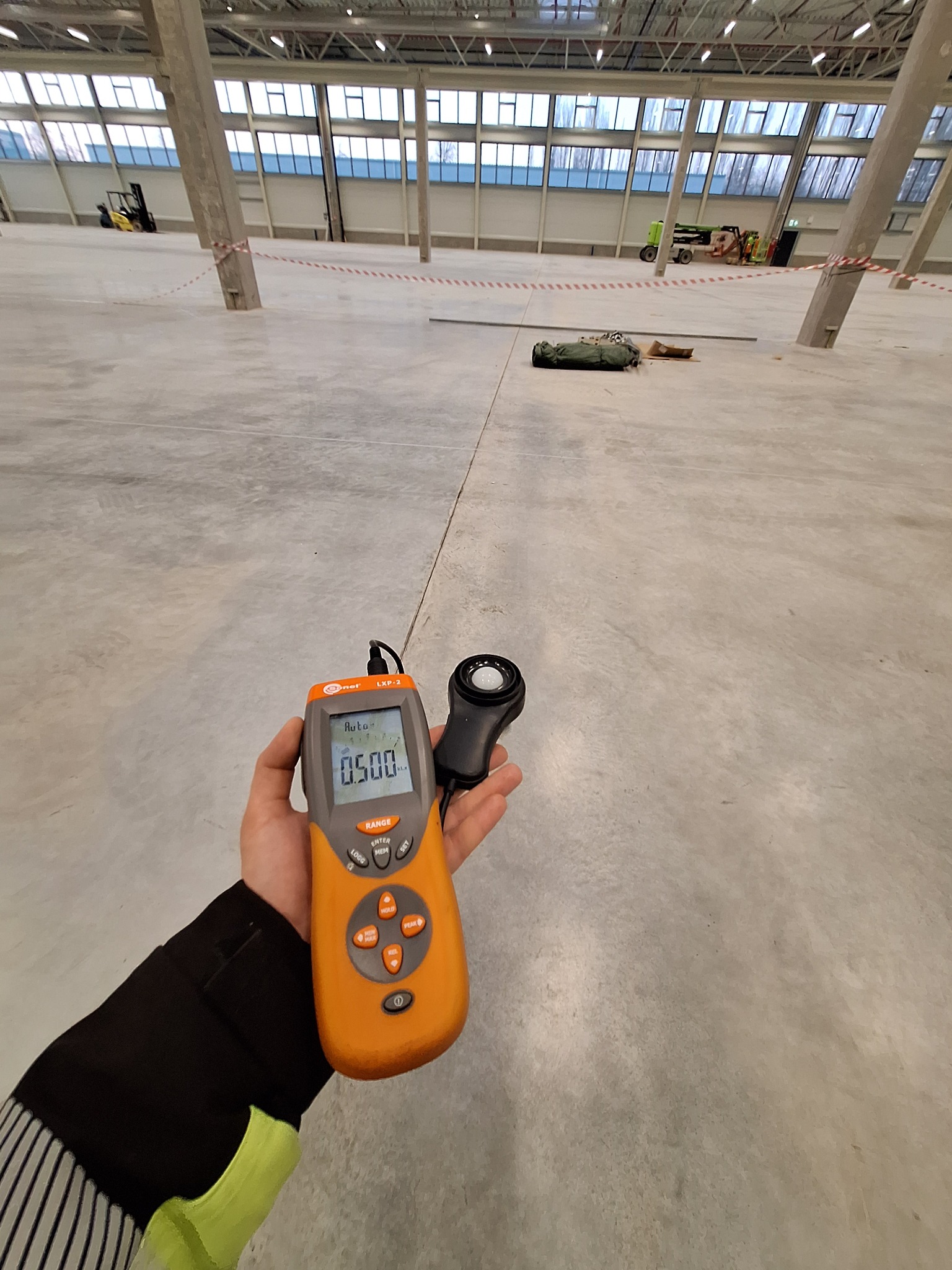 Pomiar natężenia oświetlenia luksomierzem na tle surowej posadzki w hali magazynowej z oknami i słupami. Na wyświetlaczu urządzenia widoczna wartość 0.500 lx.