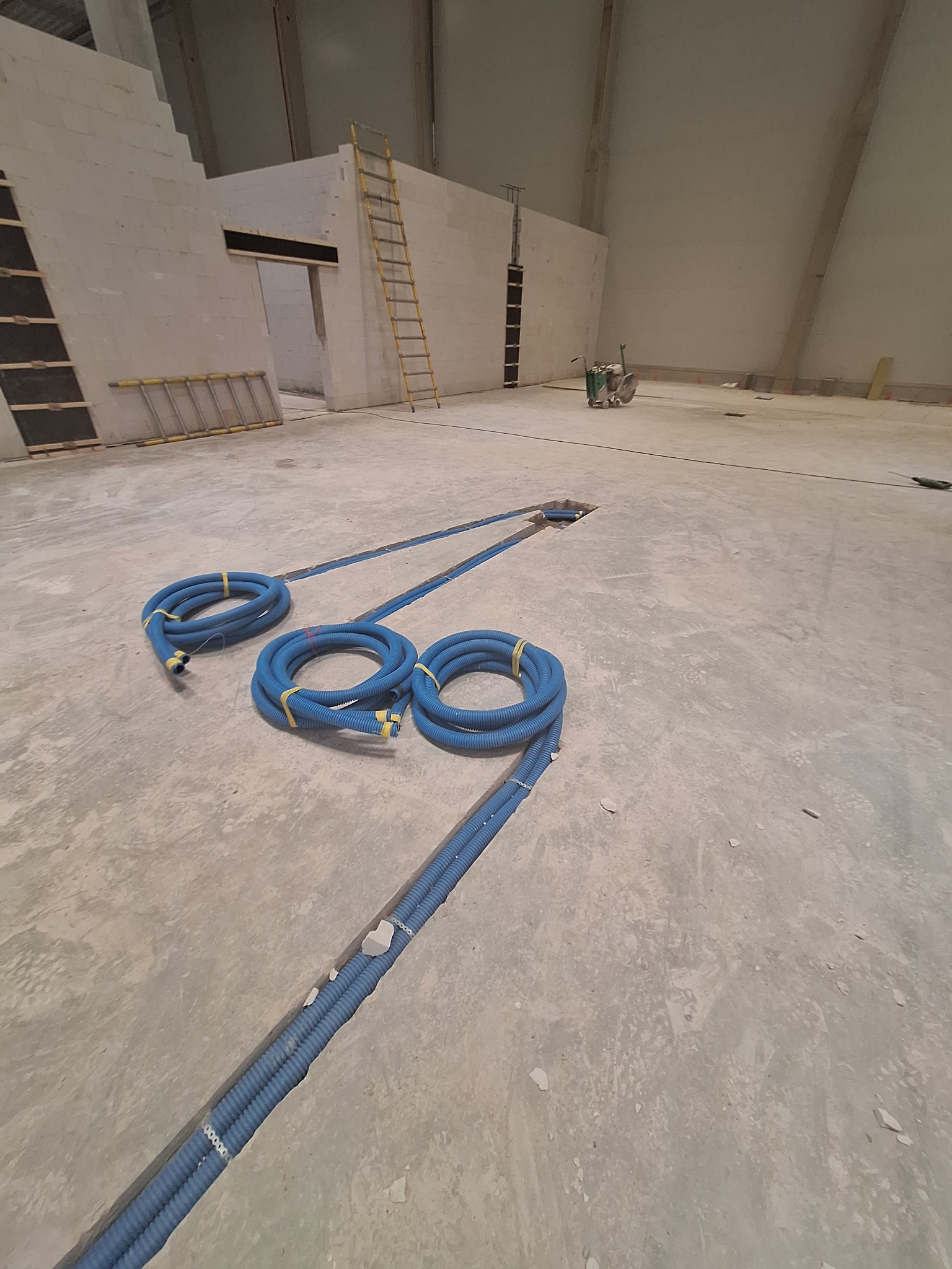 Niebieskie rury w bruzdach w betonie, układane w nowym budynku. Widoczne drabiny i maszyna do cięcia betonu. Instalacja elektryczna w trakcie realizacji.