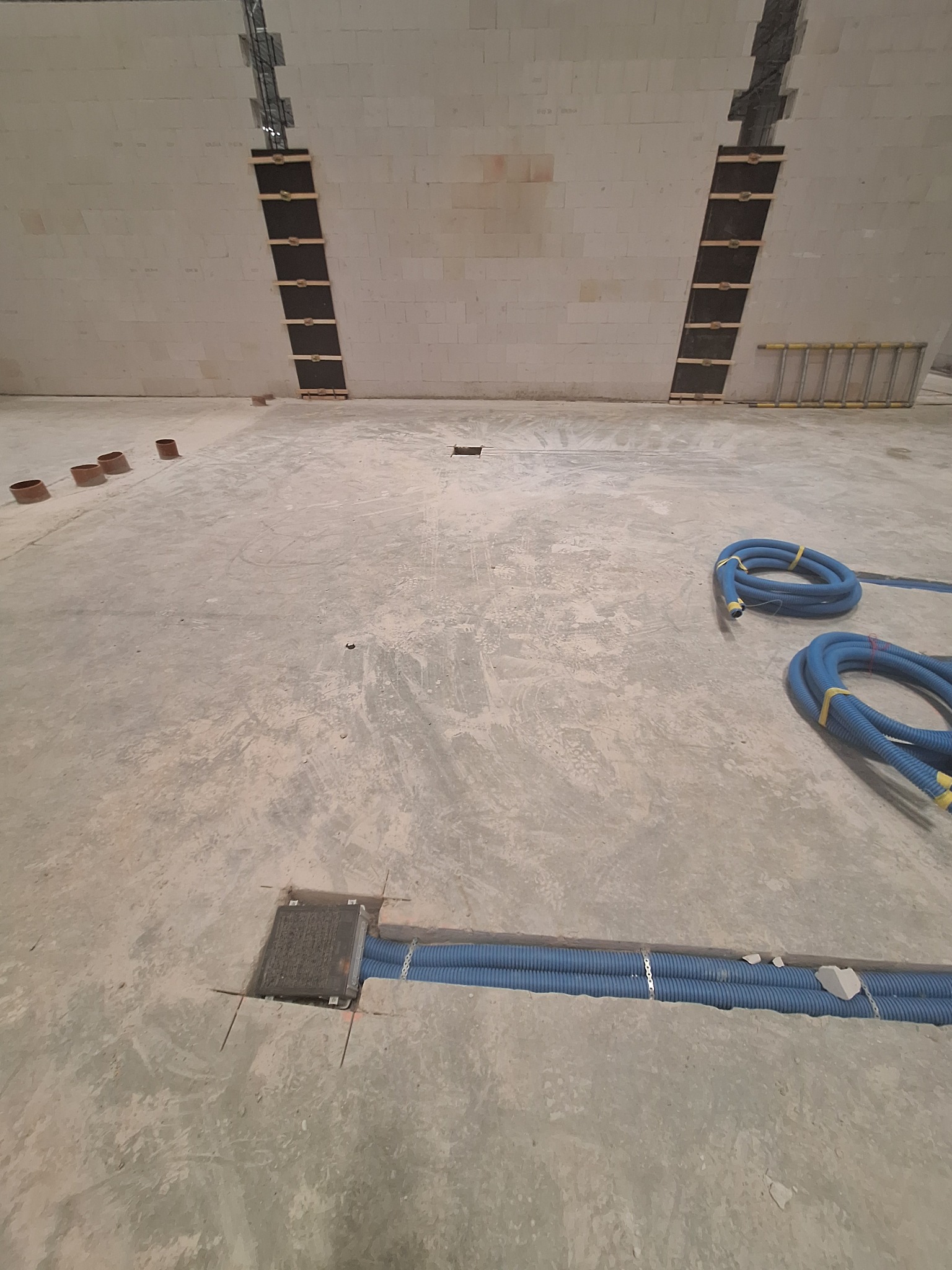 Niebieskie rury w bruzdach w podłodze, przygotowane do zalania betonem we Wrocławiu. Widoczne także pionowe kanały w ścianie i drabina w tle.