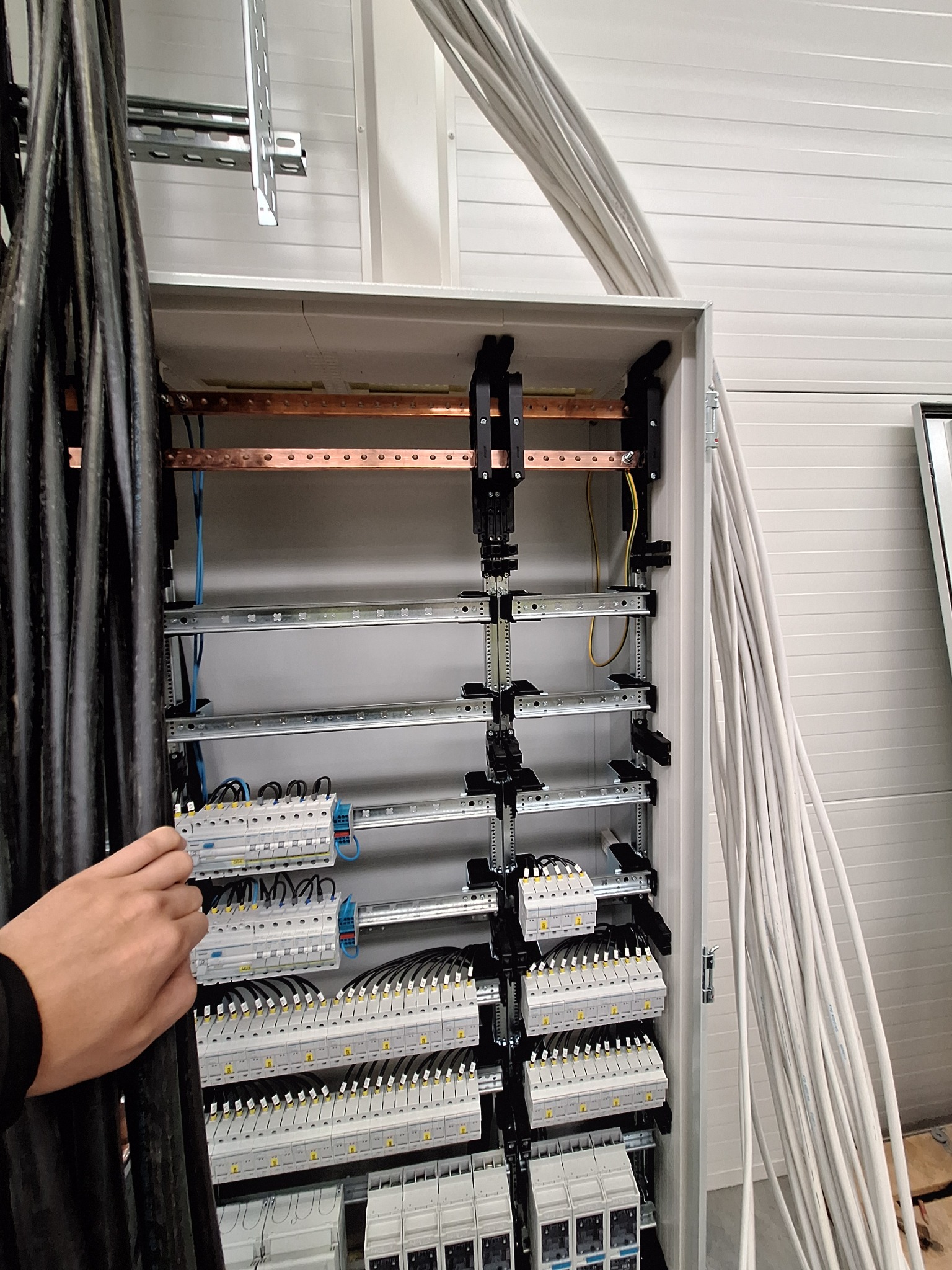 Rozdzielnia elektryczna w trakcie montażu: widoczne kable, szyny, zabezpieczenia. Ręka elektryka przytrzymuje przewody. Białe ściany w tle.