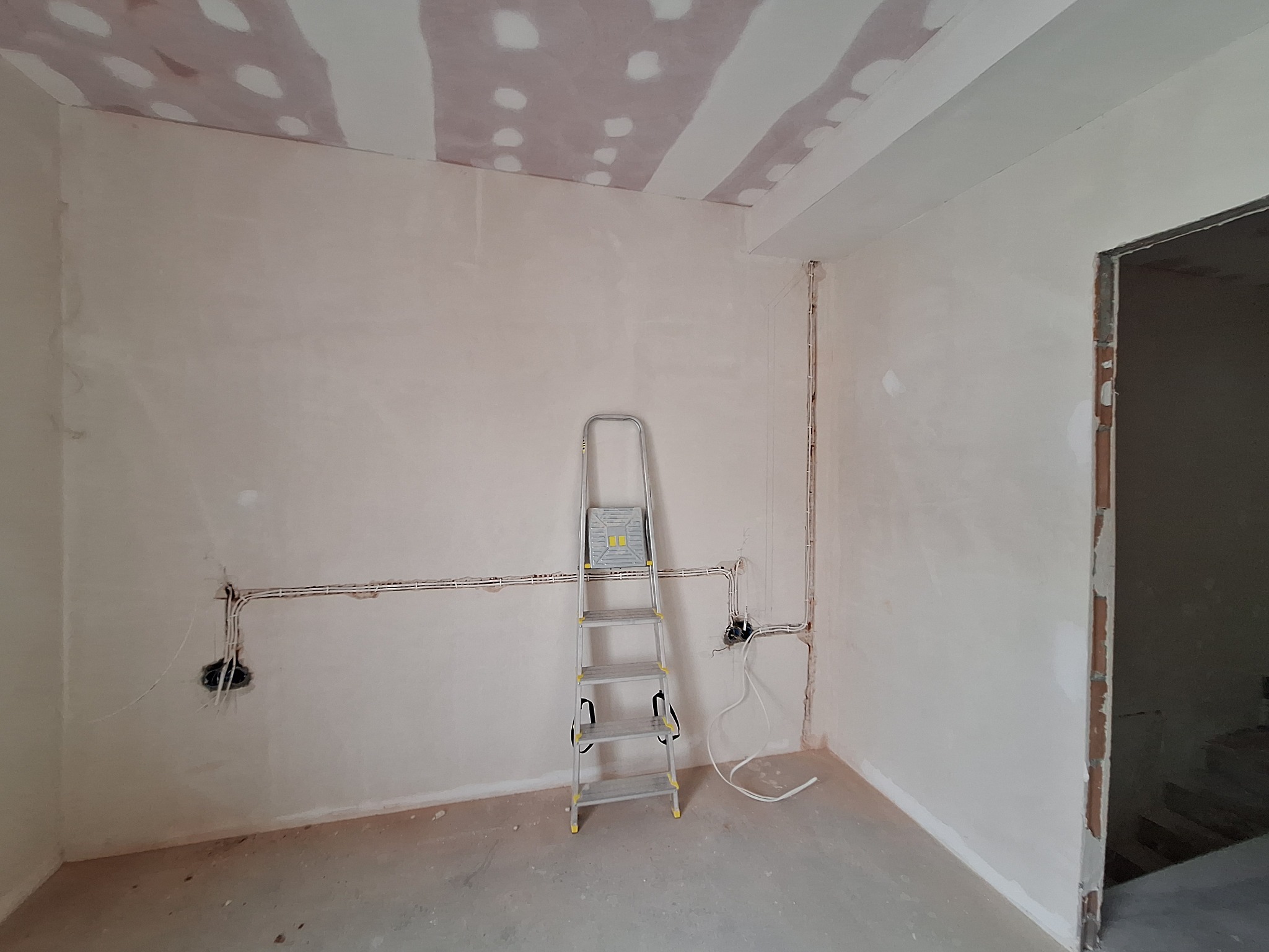 Wnętrze w trakcie remontu: ściany po gładzi, widoczne przewody elektryczne, drabina aluminiowa. Sufit z płyt gipsowych, częściowo zaszpachlowany. Otwór drzwiowy.