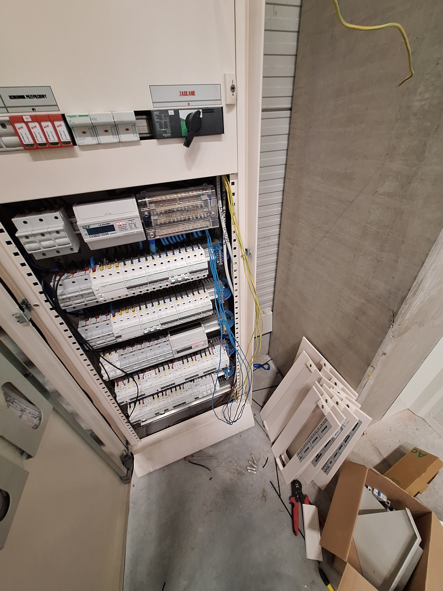 Otwarta szafa elektryczna z widocznymi zabezpieczeniami, licznikiem i okablowaniem. Na podłodze narzędzia i elementy obudowy. Widok z góry, oświetlenie sufitowe.