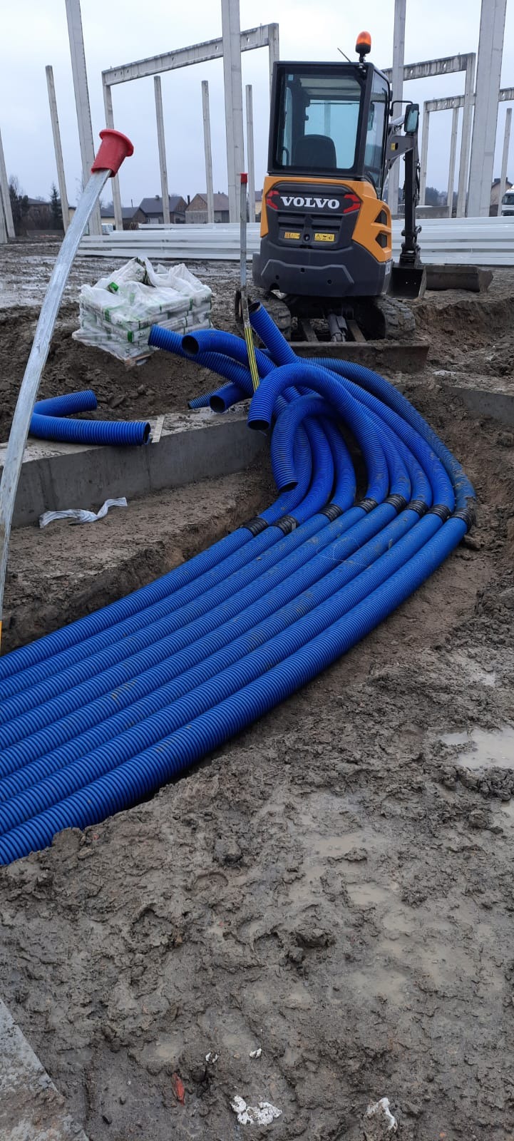 Niebieskie rury karbowane ułożone w ziemi na placu budowy, z widocznym minikoparką Volvo i elementami konstrukcji budynku w tle. Instalacja podziemna w trakcie realizacji.