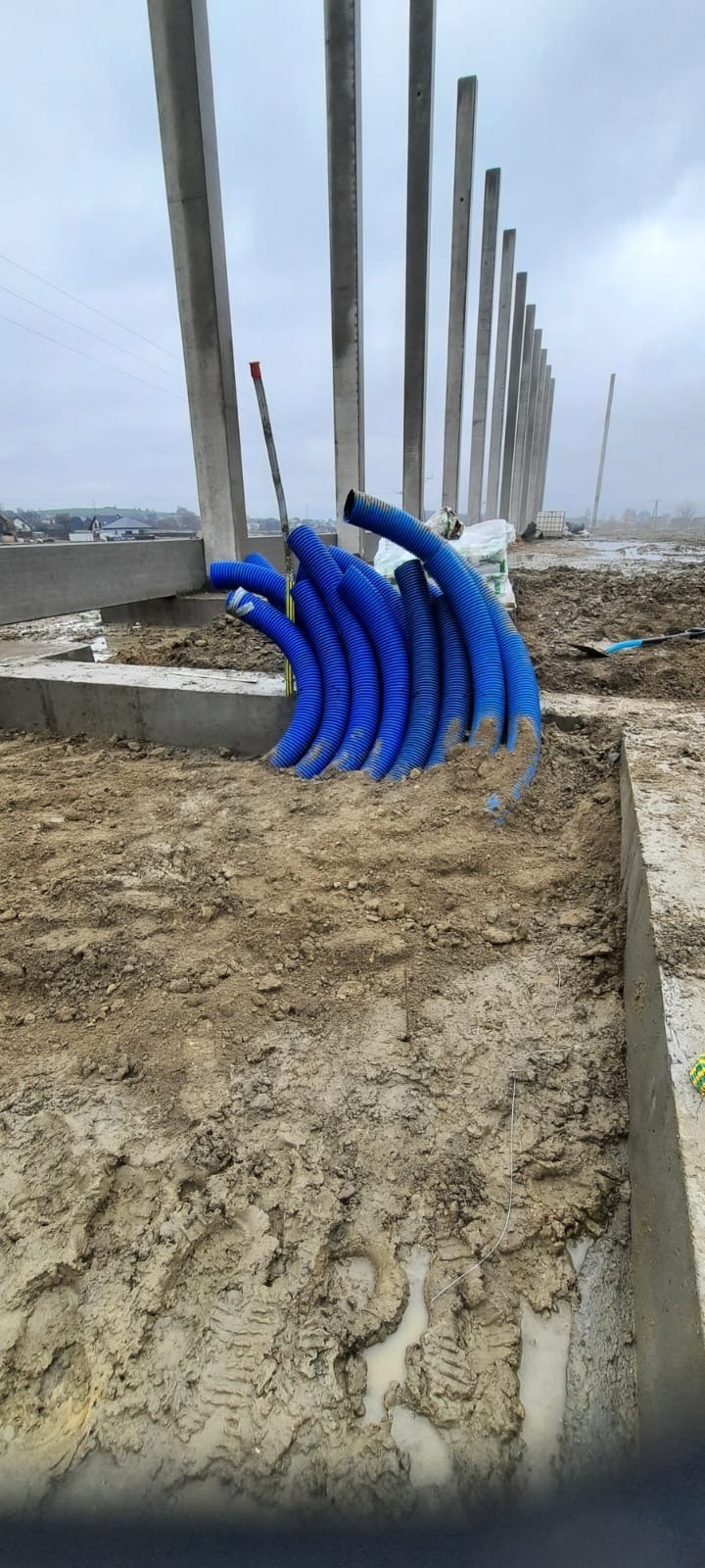 Niebieskie rury karbowane ułożone w ziemi na placu budowy, widoczne betonowe fundamenty i słupy konstrukcyjne. Pochmurne niebo w tle.