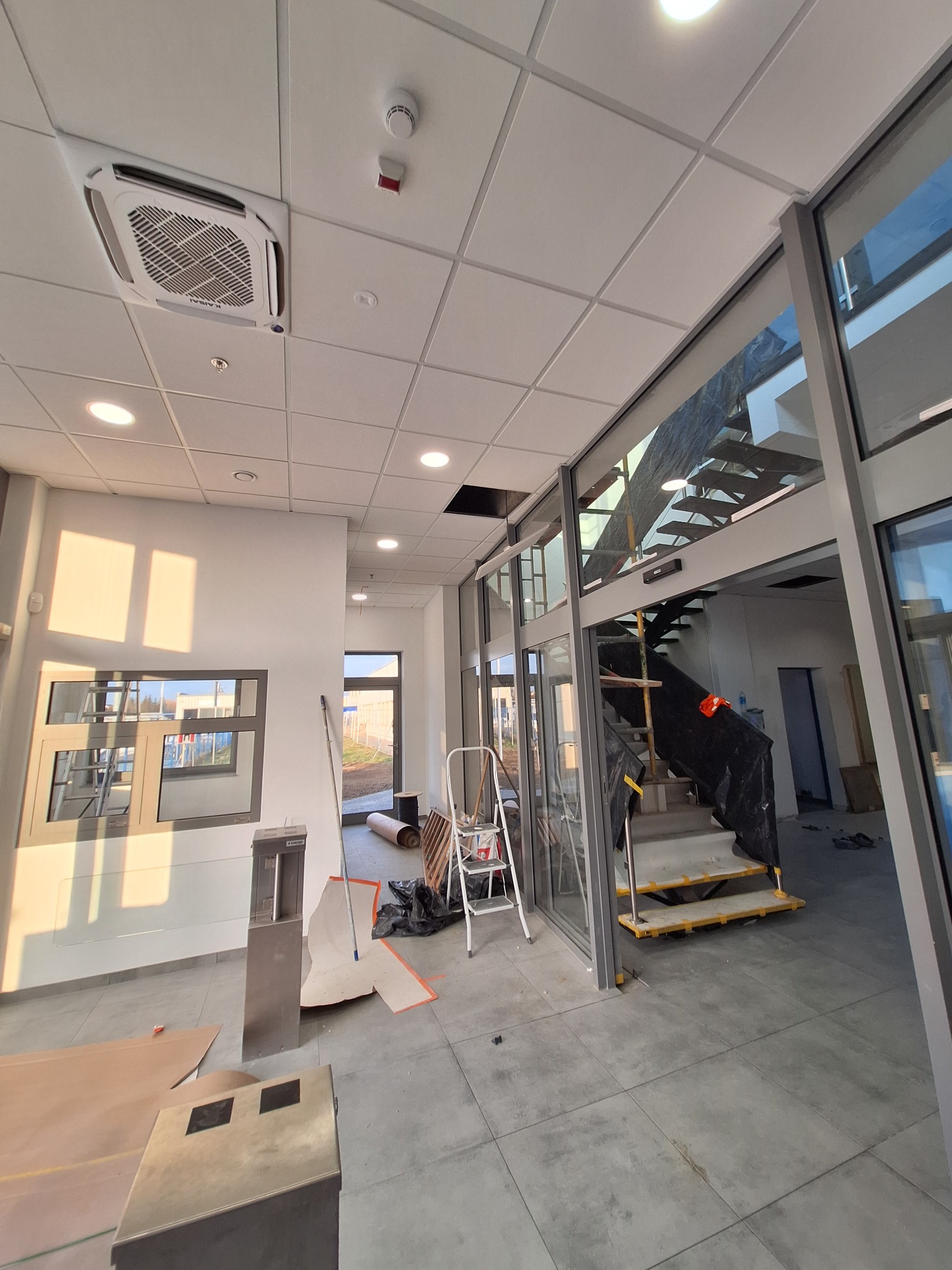 Wnętrze w trakcie remontu: widoczny sufit podwieszany z klimatyzacją kasetonową, schody zabezpieczone folią, okna i drzwi aluminiowe. W tle drabina i materiały budowlane.