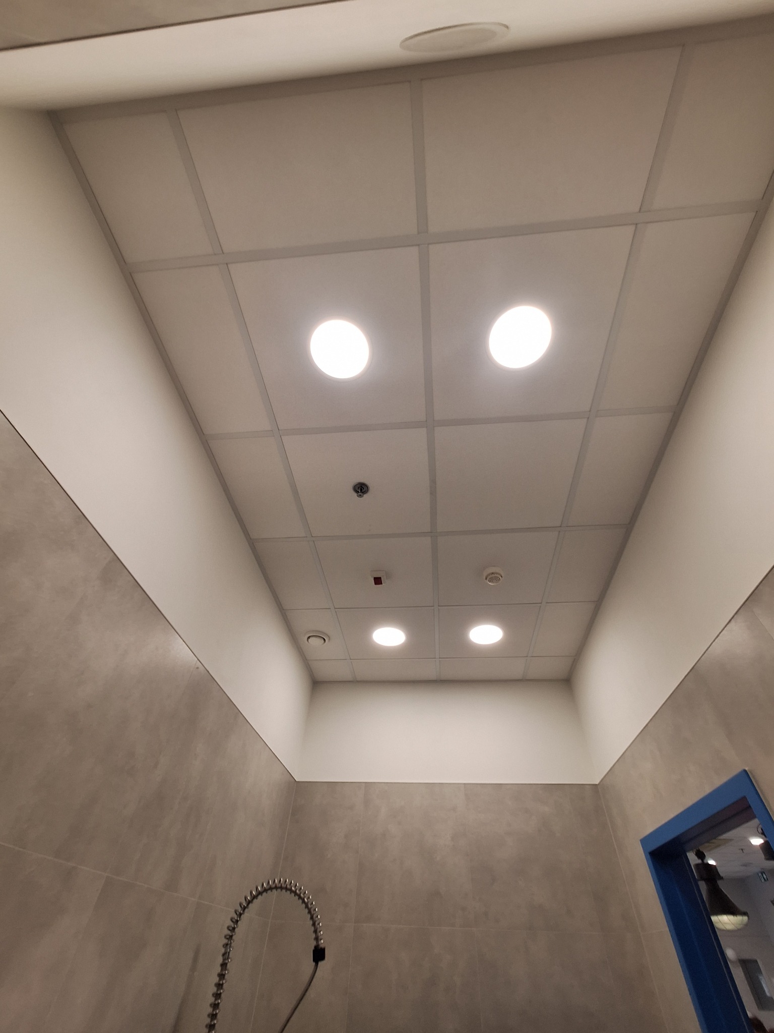 Kasetonowy sufit z oświetleniem LED i czujnikami, widok z dołu. Realizacja we Wrocławiu. Szara ściana i fragment niebieskiej ramy drzwi w tle.