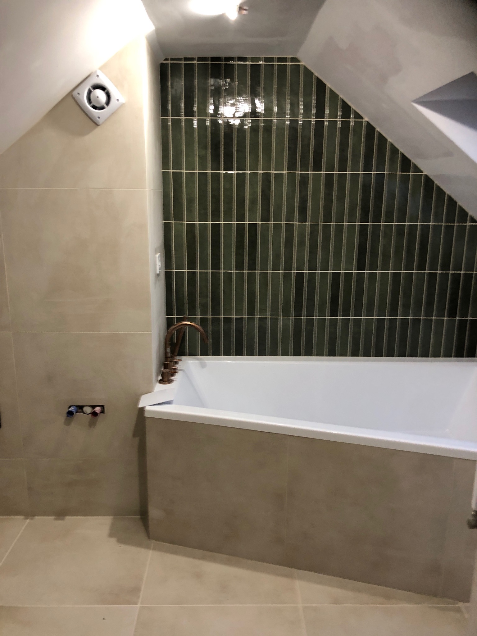 Wykończona łazienka na poddaszu z wanną, beżowymi płytkami i pionowymi, ciemnozielonymi płytkami na ścianie pod skosem. Widoczna miedziana bateria i wentylator.