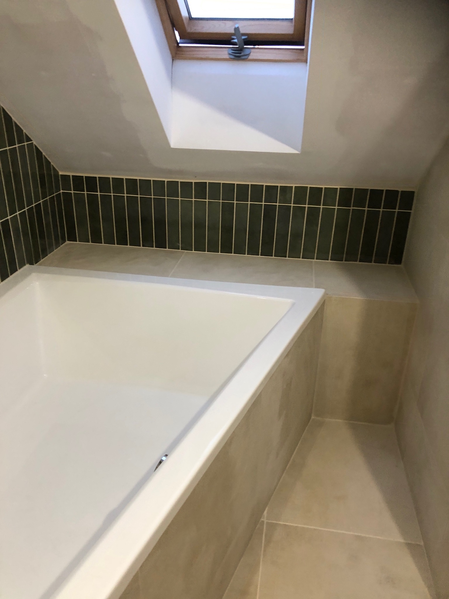 Wykończona łazienka na poddaszu z białą wanną, obłożona ciemnozielonymi, pionowymi płytkami i jasnymi płytkami podłogowymi. Widoczne okno dachowe.