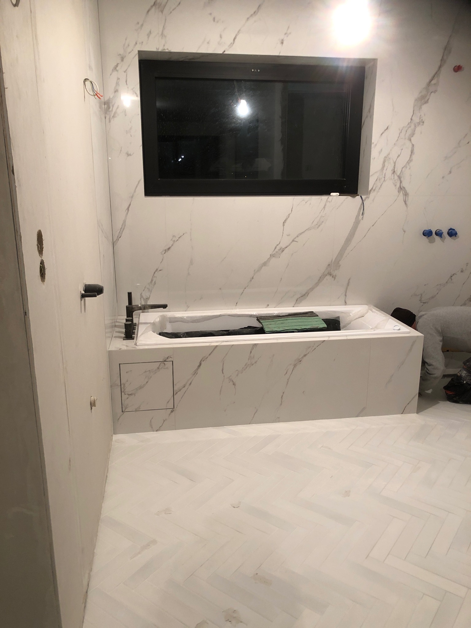 Nowoczesna łazienka z wanną obudowaną płytkami imitującymi marmur, czarną armaturą i oknem. Na podłodze biała jodełka. Widoczny fragment osoby podczas prac wykończeniowych.