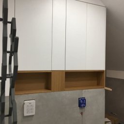 W&T Stolarstwo - Biała szafa wnękowa z drewnianymi półkami w nowoczesnym wnętrzu, widok z dołu, oświetlenie dzienne, elementy instalacji na ścianie.