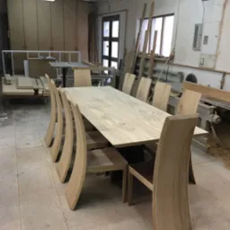 Długi, jasny stół z krzesłami o zakrzywionych oparciach, wykonany z surowego drewna, stoi w warsztacie stolarskim. W tle widoczne narzędzia i materiały.