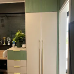 Nowoczesna szafa z frezowanymi frontami w odcieniach zieleni i beżu, z długimi, smukłymi uchwytami. Na szafce dekoracje, w tle okno z żaluzjami.