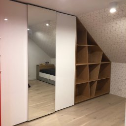 W&T Stolarstwo - Biała szafa wnękowa z lustrem i drewnianym regałem na poddaszu, z wzorzystą tapetą na ścianie i jasną podłogą, oświetlona lampami sufitowymi.