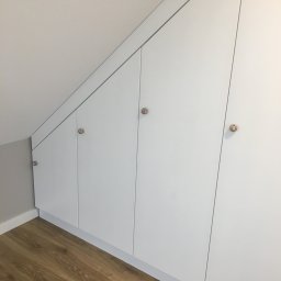 W&T Stolarstwo - Biała szafa wnękowa na poddaszu z drewnianą podłogą i ozdobnymi, kolorowymi gałkami. Minimalistyczny design, dopasowanie do skosu dachu.