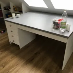 Biało-szare biurko z szufladami i blatem roboczym, stojące na drewnianej podłodze. Na blacie pojemnik z akcesoriami, szklanka, butelka i rolka papieru.