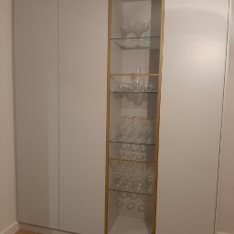 W&T Stolarstwo - Biała szafa wnękowa z przeszkloną witryną o złotej ramie, eksponująca kieliszki na szklanych półkach. Minimalistyczny design, parkiet na podłodze.
