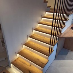W&T Stolarstwo - Drewniane schody z podświetleniem LED, czarne metalowe barierki. Naturalne drewno z widocznymi słojami. Minimalistyczny design, białe ściany.