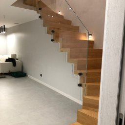 W&T Stolarstwo - Nowoczesne schody drewniane z balustradą ze szkła, oświetleniem punktowym i minimalistycznym designem w jasnym wnętrzu. Widok z dołu, podkreślający detale konstrukcji.