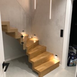 W&T Stolarstwo - Nowoczesne, drewniane schody wiszące z podświetleniem LED i szklaną balustradą. Minimalistyczny design, jasne drewno, szare ściany z dekoracyjnym tynkiem.