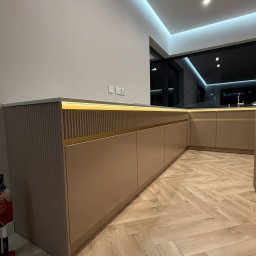 Beżowa zabudowa kuchenna z ryflowanymi frontami i podświetleniem LED, ułożona w kształcie litery L, na parkiecie w jodełkę. Nowoczesny design z nutą elegancji.