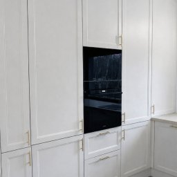 Biała zabudowa kuchenna z frezowanymi frontami, złotymi uchwytami i wbudowanym czarnym piekarnikiem. Minimalistyczny design, jasna podłoga.