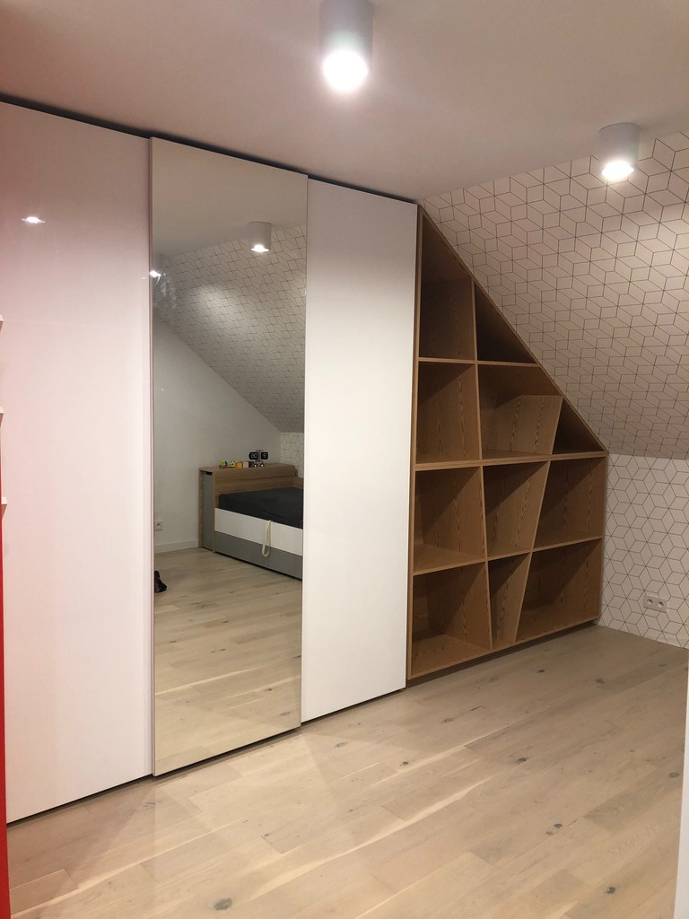 Biała szafa wnękowa z lustrem i drewnianym regałem na poddaszu, z wzorzystą tapetą na ścianie i jasną podłogą, oświetlona lampami sufitowymi.