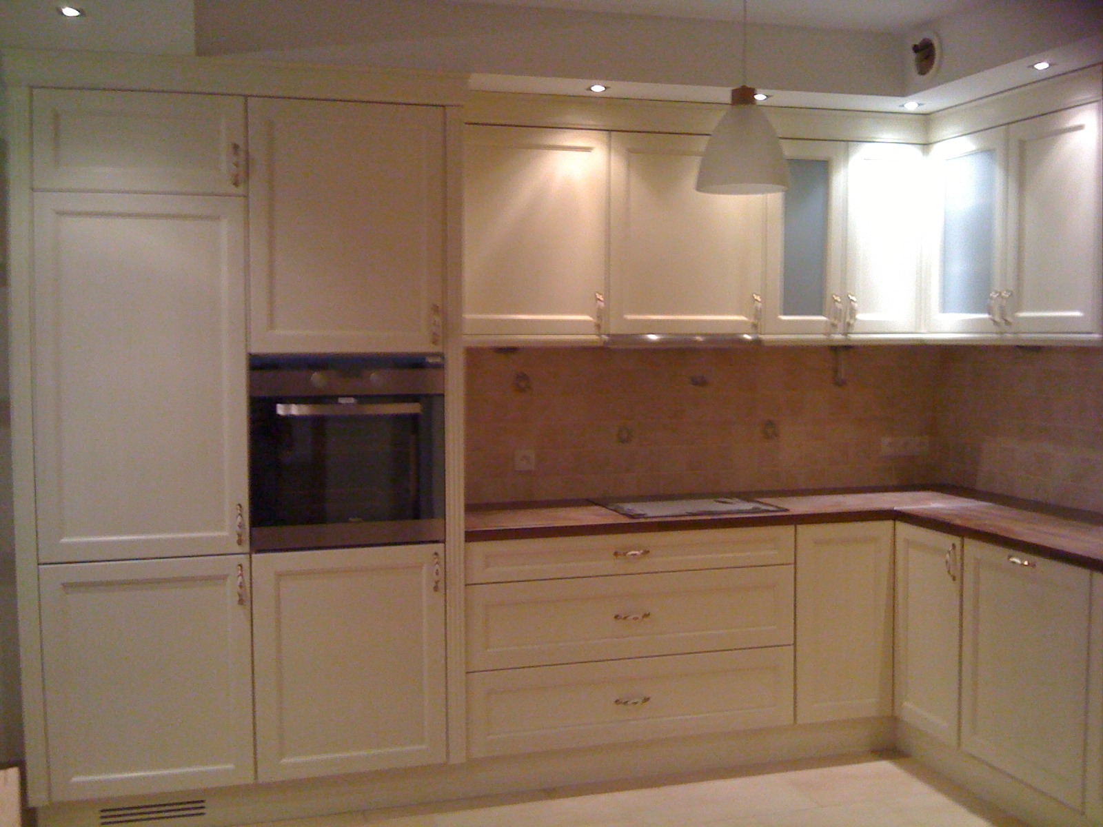 Jasna, klasyczna zabudowa kuchenna z drewnianymi frontami, wbudowanym piekarnikiem i oświetleniem punktowym. Uwagę zwracają frezowane fronty szafek i drewniany blat.