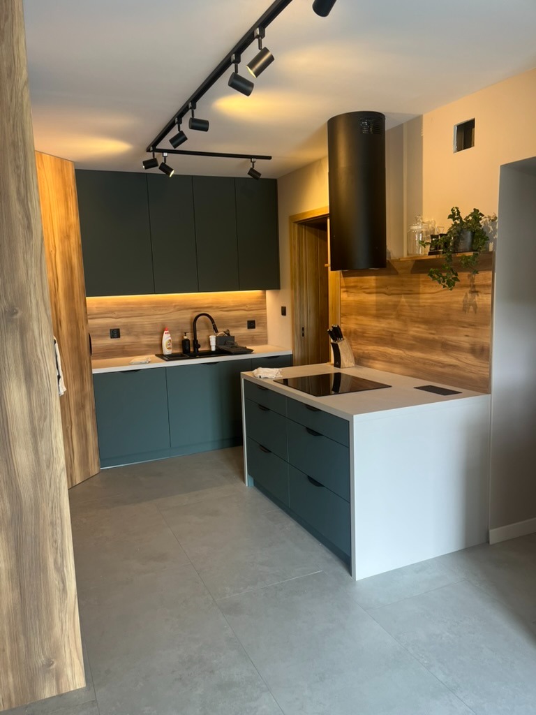 Nowoczesna kuchnia z wyspą: ciemnozielone szafki, drewniane akcenty, czarny okap i oświetlenie punktowe. Minimalistyczny design z blatem z białego konglomeratu.