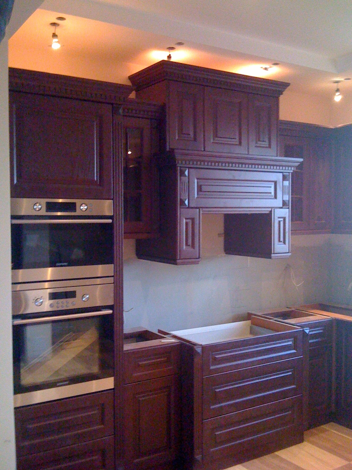 Ciemnobrązowe, klasyczne meble kuchenne w trakcie montażu: szafki wiszące, zabudowa piekarnika i okap z dekoracyjnymi frezami. Widoczne niedokończone szuflady.