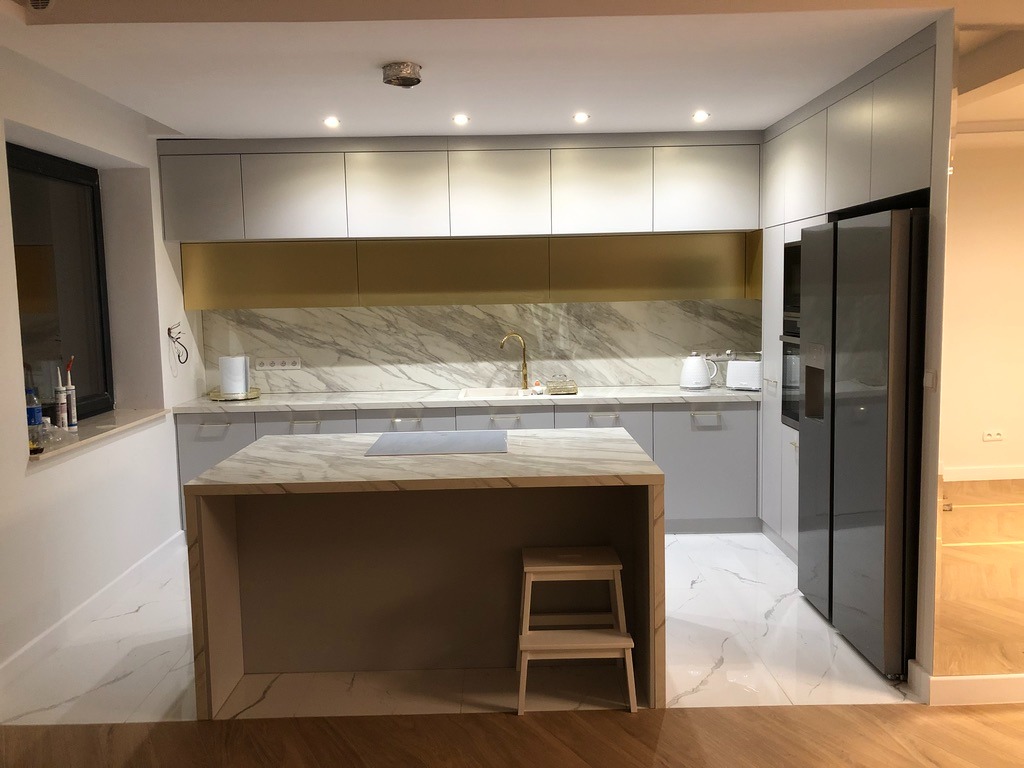 Nowoczesna kuchnia z wyspą i marmurowym blatem. Szare szafki, złote akcenty na ścianie, oświetlenie punktowe. Minimalistyczny design, drewniana podłoga.