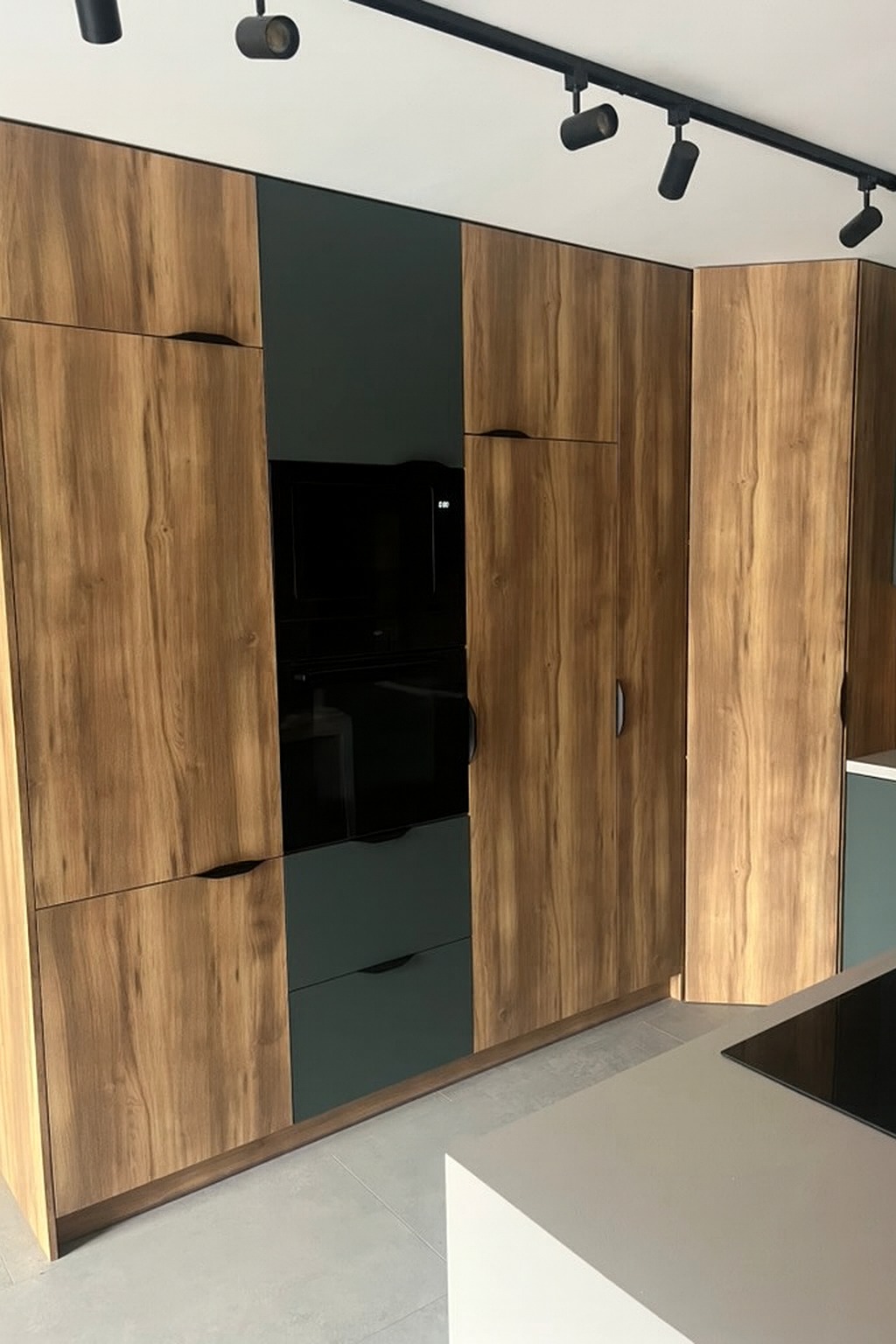 Nowoczesna zabudowa kuchenna: drewnopodobne fronty, grafitowe akcenty, minimalistyczne uchwyty. Widoczna część wyspy kuchennej i oświetlenie szynowe.