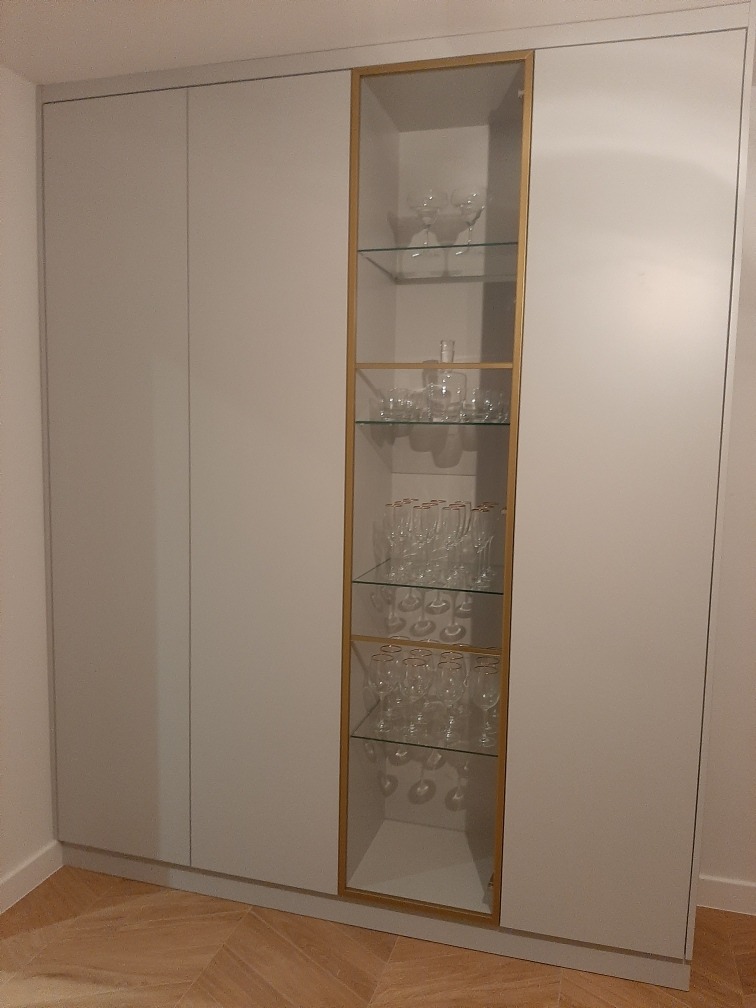 Biała szafa wnękowa z przeszkloną witryną o złotej ramie, eksponująca kieliszki na szklanych półkach. Minimalistyczny design, parkiet na podłodze.