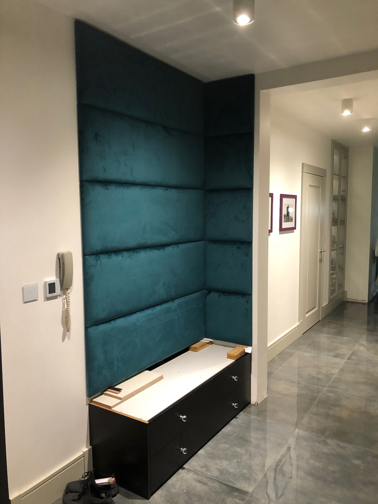 Wnęka z siedziskiem i tapicerowanym panelem ściennym w odcieniach turkusu. Widoczne wykończenie szuflad i elementy montażowe. Nowoczesny design korytarza.