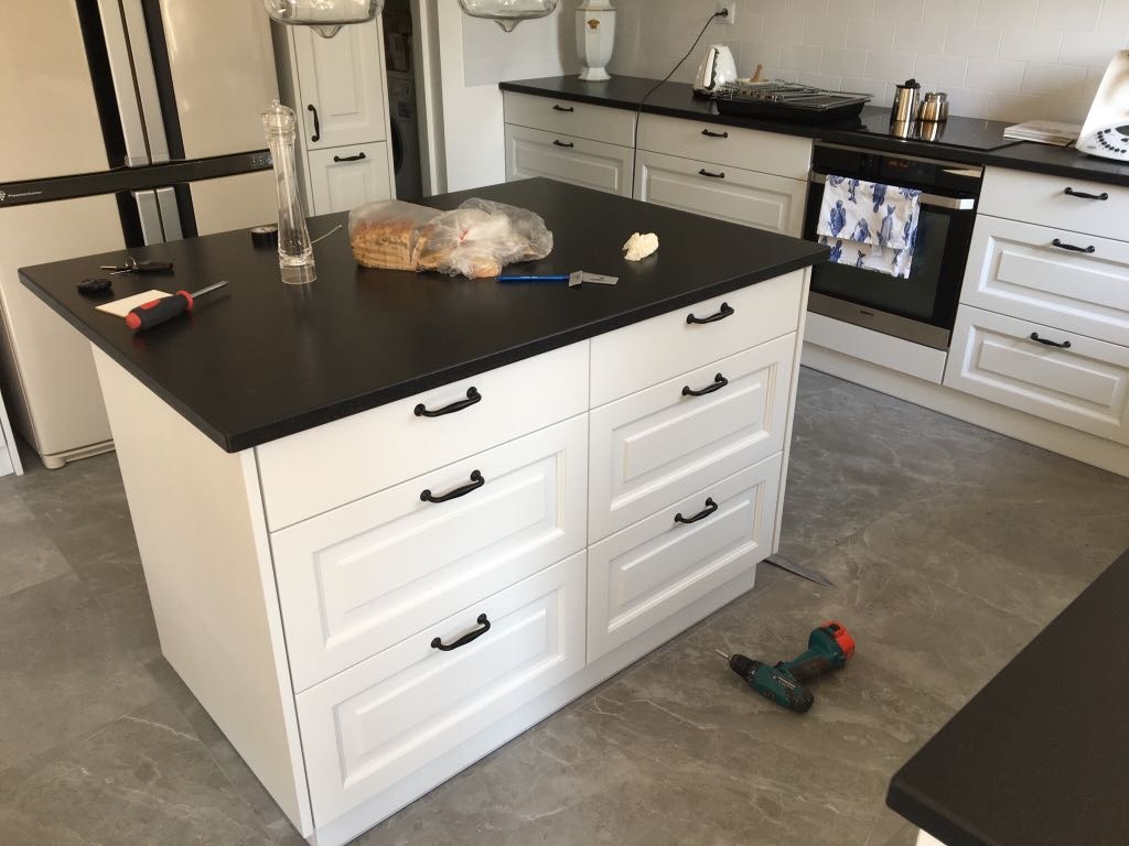 Wyspa kuchenna z białymi frontami szuflad i czarnym blatem, narzędzia na blacie i podłodze, w tle zabudowa kuchenna z piekarnikiem i lodówką.