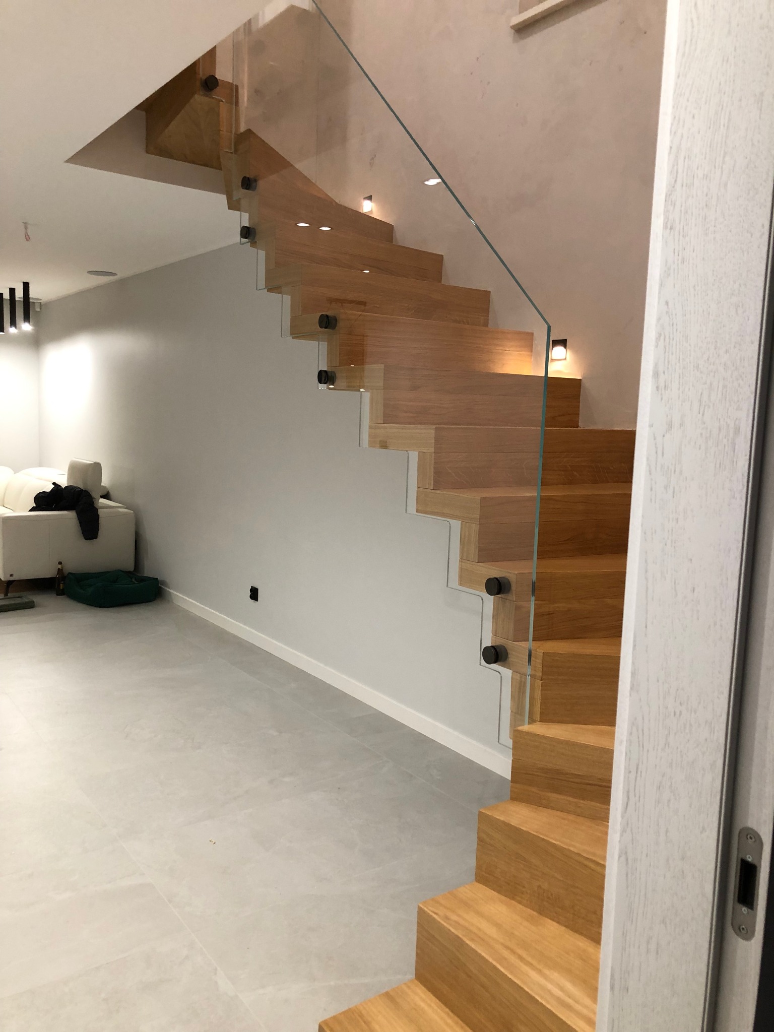 Nowoczesne schody drewniane z balustradą ze szkła, oświetleniem punktowym i minimalistycznym designem w jasnym wnętrzu. Widok z dołu, podkreślający detale konstrukcji.