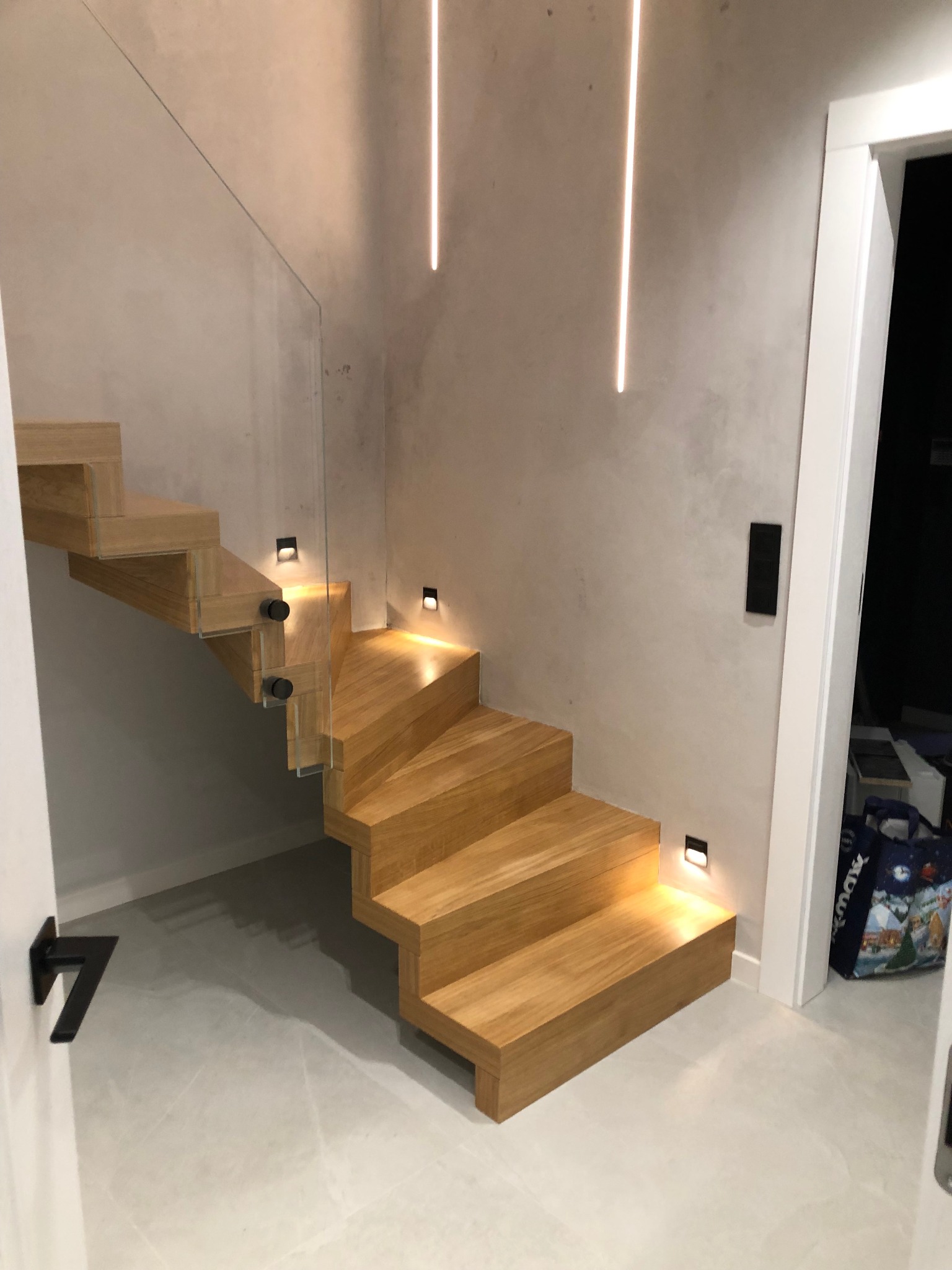 Nowoczesne, drewniane schody wiszące z podświetleniem LED i szklaną balustradą. Minimalistyczny design, jasne drewno, szare ściany z dekoracyjnym tynkiem.