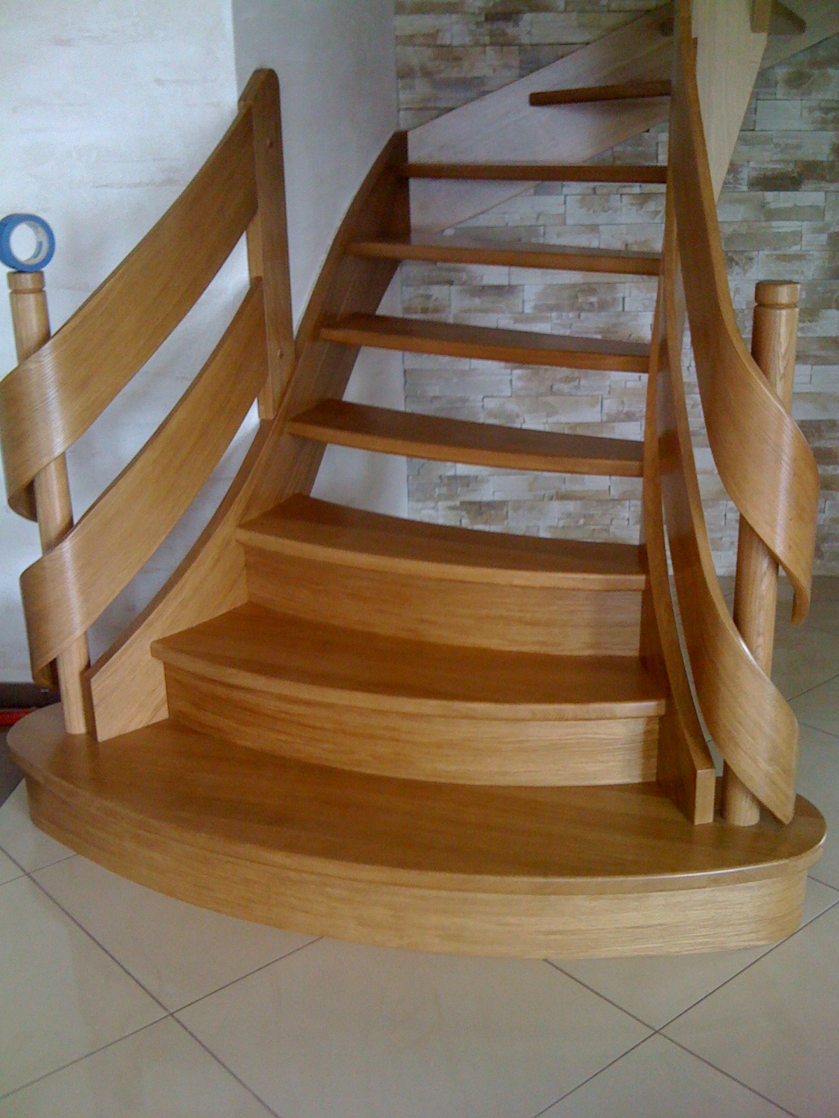 Drewniane schody z jasnego drewna z zakręconą balustradą, widok z dołu. Stopnie zaokrąglone na krawędziach, tło stanowi ściana z imitacją kamienia.