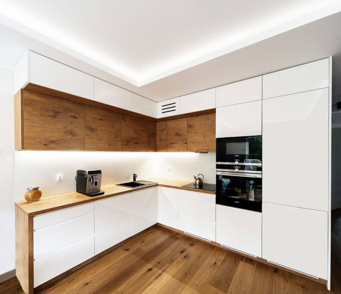 Nowoczesna kuchnia w kształcie litery L z białymi, lakierowanymi frontami i drewnianymi szafkami górnymi. Na blacie ekspres do kawy i szklany słoik. Parkiet na podłodze.