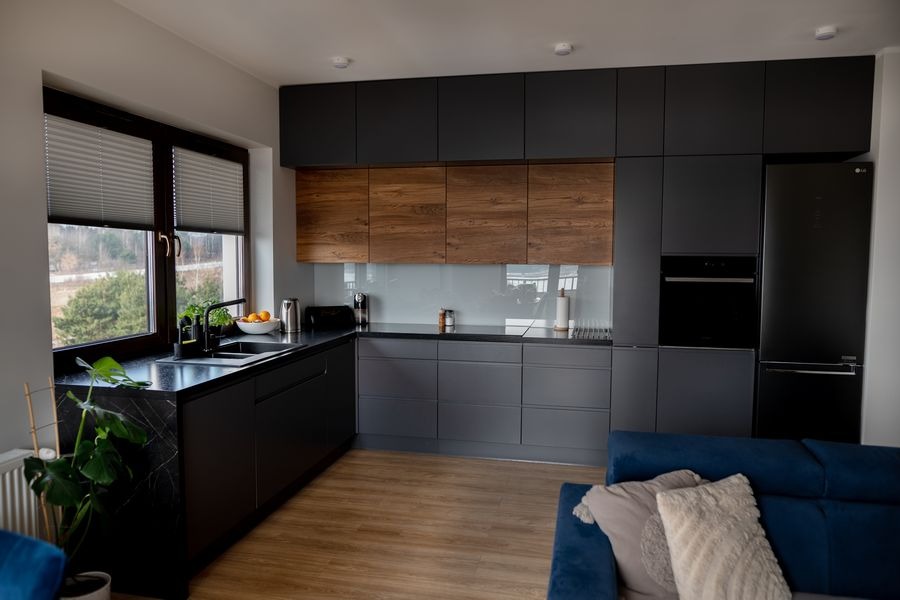 Nowoczesna kuchnia z ciemnymi, matowymi frontami i drewnianymi akcentami. Marmurowy blat przy oknie z widokiem na zieleń. Minimalistyczny design z elementami dekoracyjnymi.