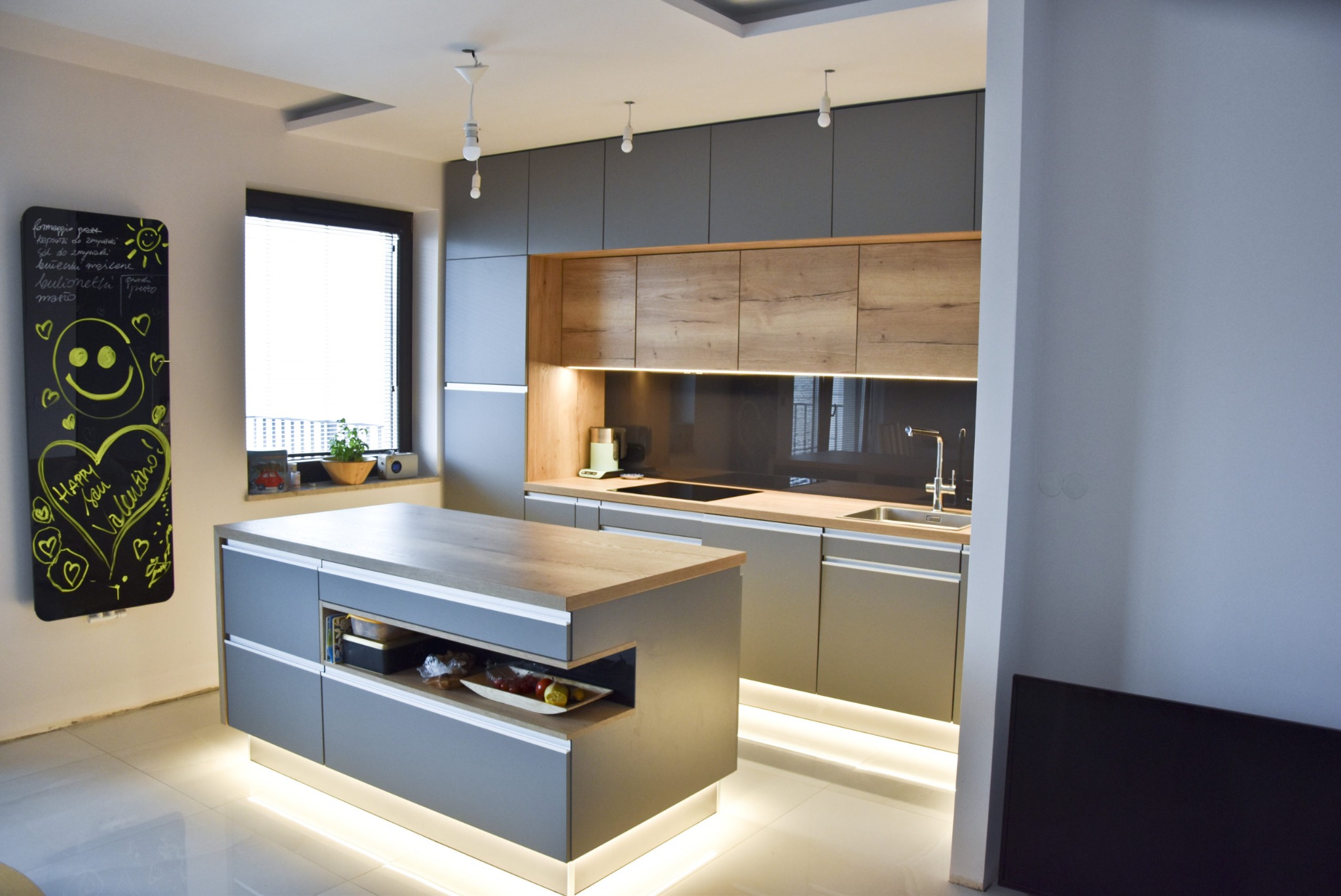 Nowoczesna kuchnia z szarymi szafkami i drewnianymi akcentami, oświetleniem LED pod wyspą. Na ścianie czarna tablica z rysunkami. Minimalistyczny design.