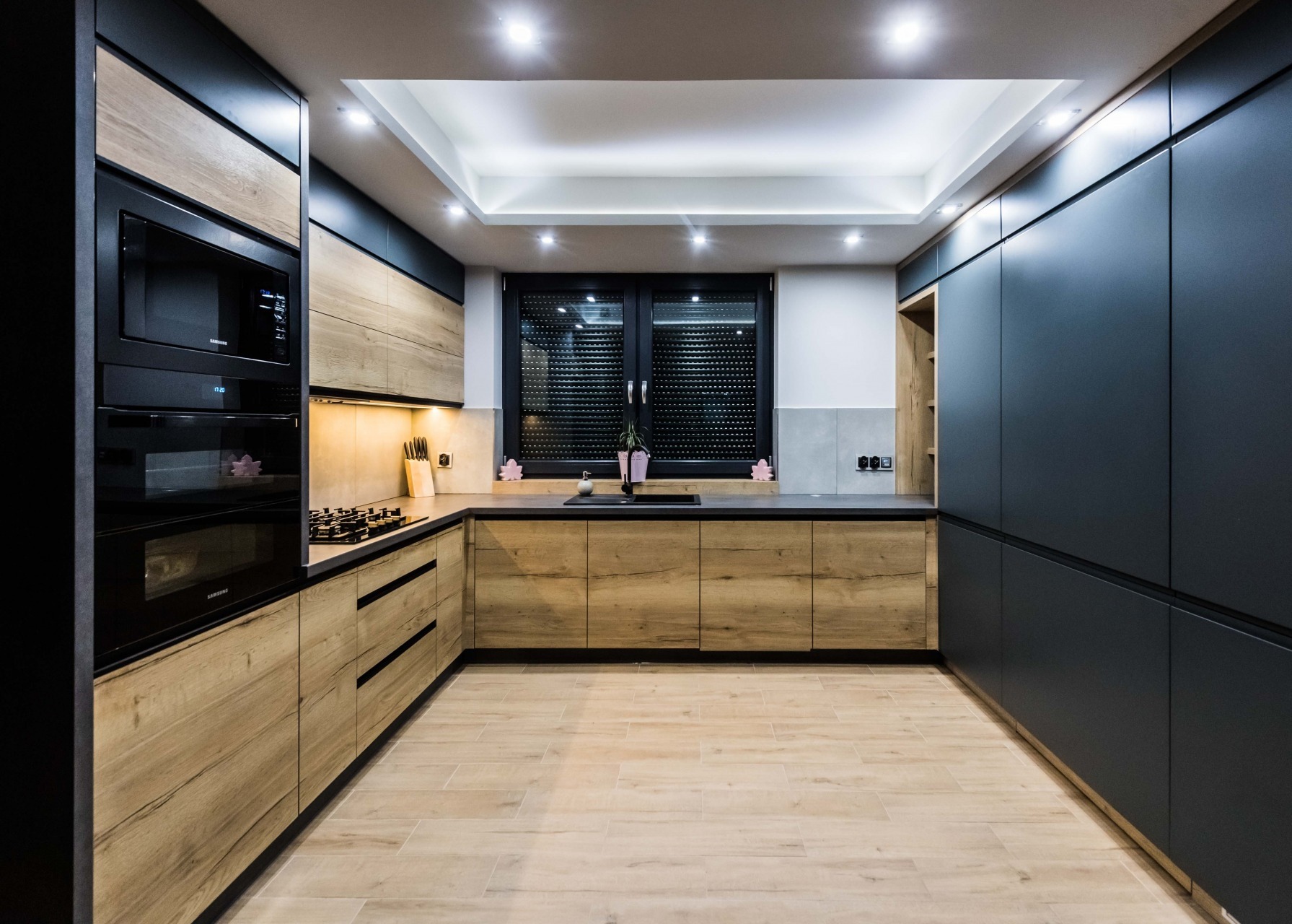 Nowoczesna kuchnia na wymiar z drewnianymi frontami i czarnymi szafkami, oświetleniem LED i płytkami imitującymi drewno na podłodze. Widok na okno z roletami.