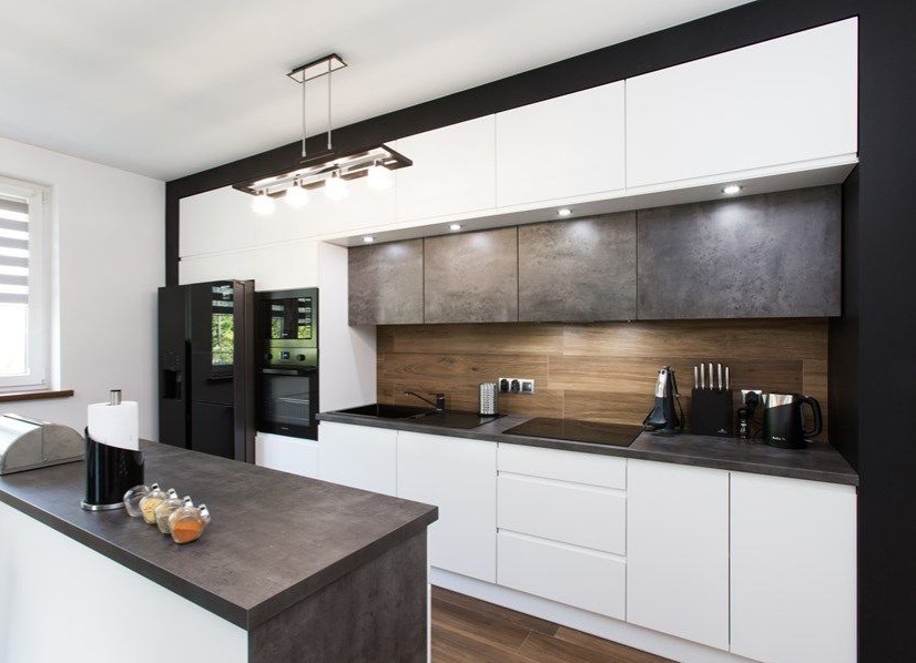 Nowoczesna kuchnia z białymi szafkami, szarym blatem i drewnianym panelem. Oświetlenie LED i czarne akcenty dodają elegancji. Widoczny sprzęt AGD.