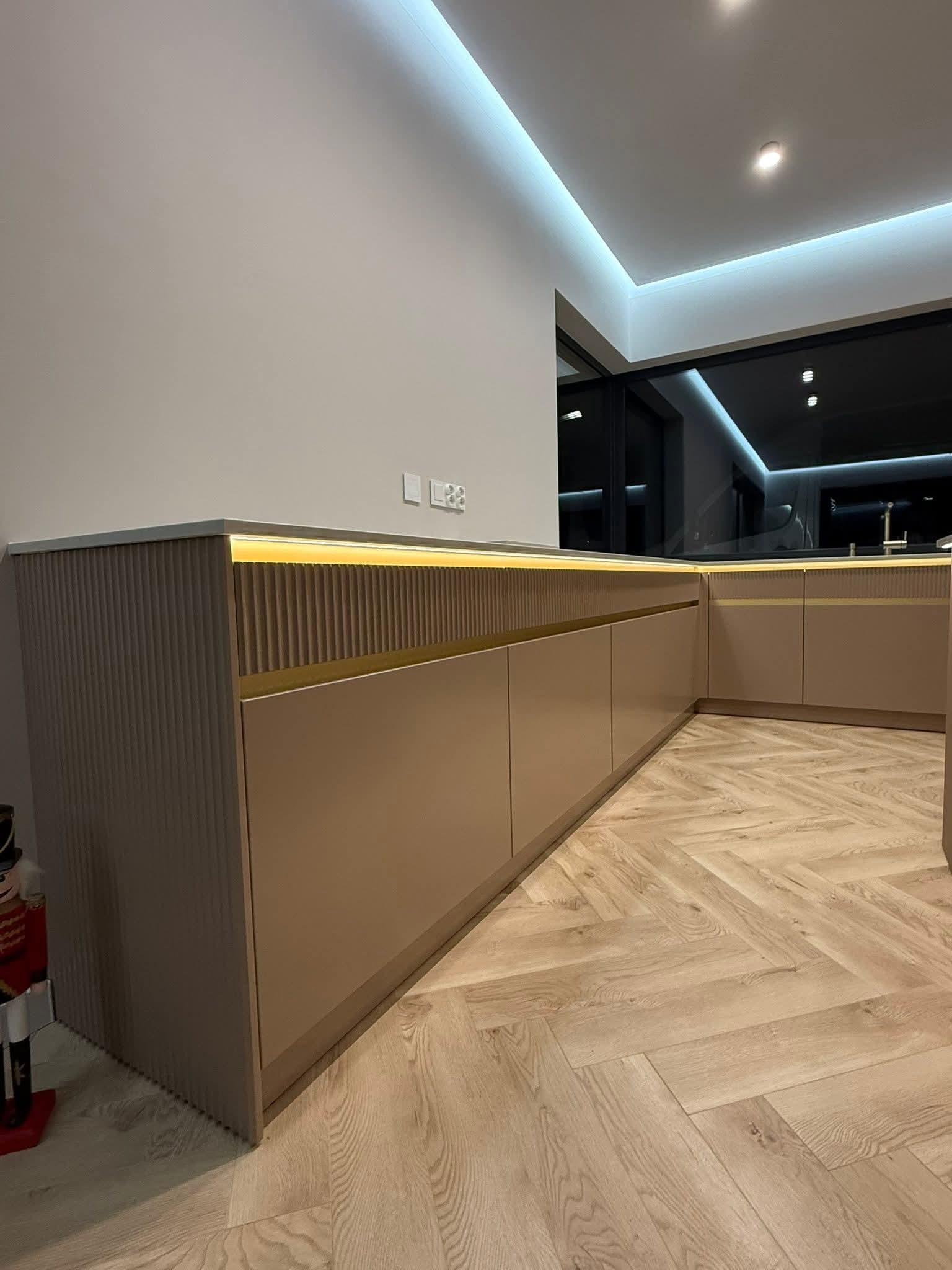 Beżowa zabudowa kuchenna z ryflowanymi frontami i podświetleniem LED, ułożona w kształcie litery L, na parkiecie w jodełkę. Nowoczesny design z nutą elegancji.