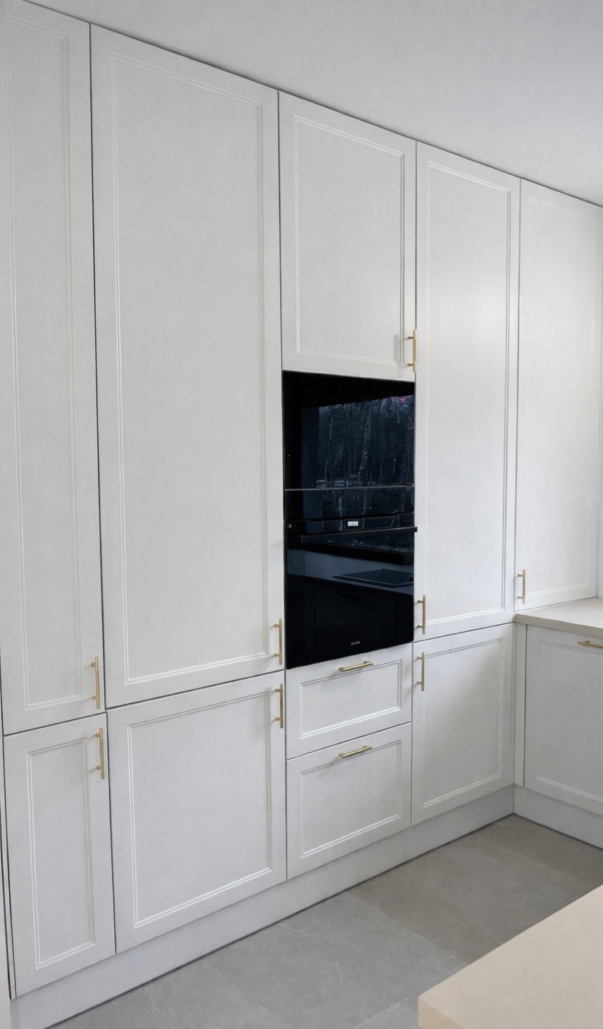 Biała zabudowa kuchenna z frezowanymi frontami, złotymi uchwytami i wbudowanym czarnym piekarnikiem. Minimalistyczny design, jasna podłoga.