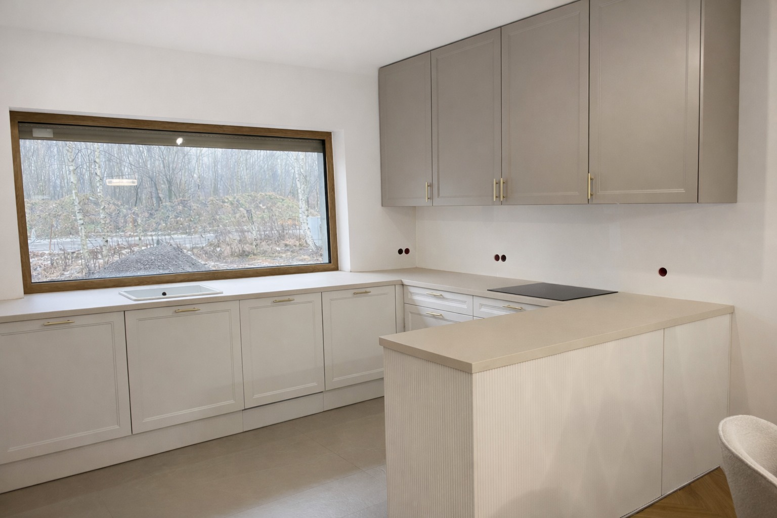 Jasna kuchnia z beżowymi frontami i złotymi uchwytami, widok na okno z blatem. Minimalistyczny design, nowoczesne wykończenie, płyta indukcyjna na blacie.