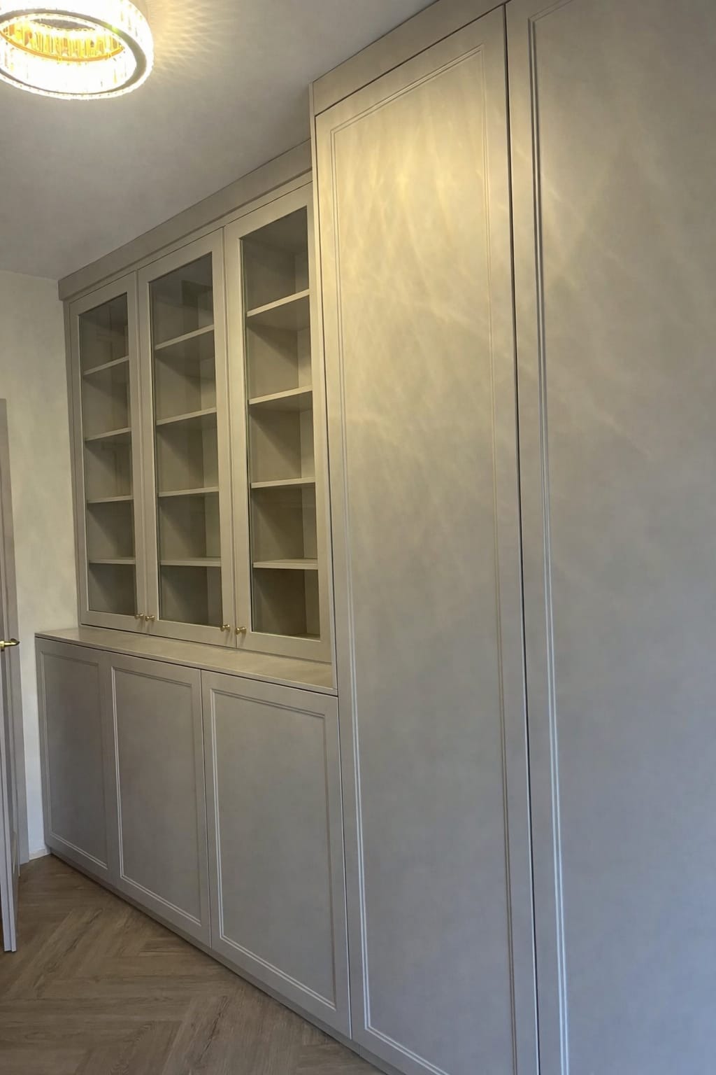 Jasnoszara zabudowa meblowa z przeszklonymi witrynami i pełnymi szafkami, oświetlona lampą sufitową. Parkiet ułożony w jodełkę.