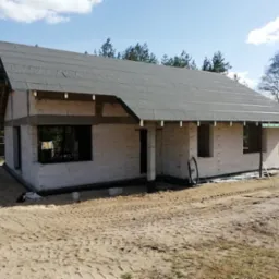 Stan surowy domu murowanego z białego bloczka, z czarną membraną na dachu w Iłowo-Osada. Widoczne otwory okienne i drzwiowe, teren piaszczysty, drzewa w tle.