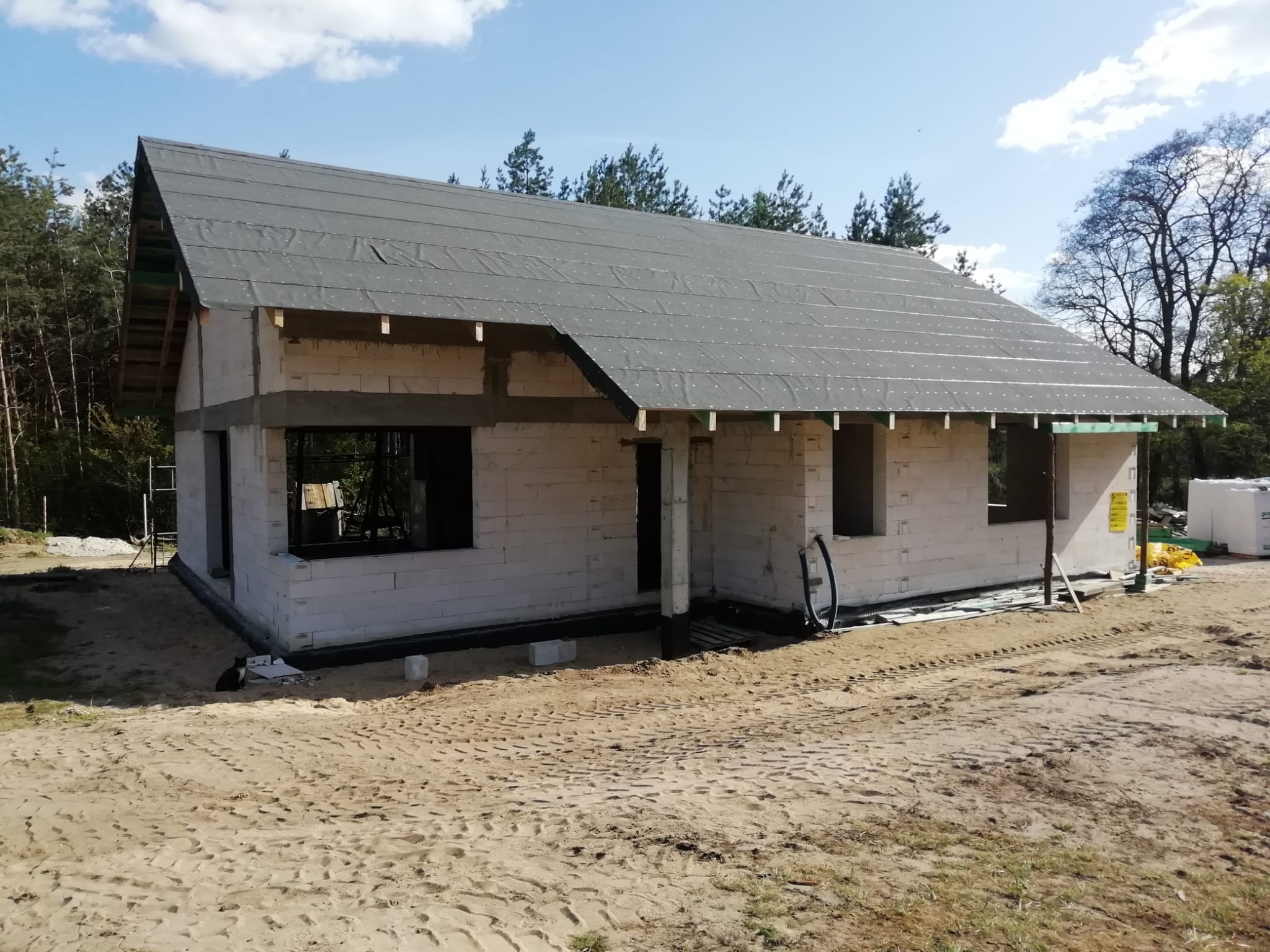 Stan surowy domu murowanego z białego bloczka, z czarną membraną na dachu w Iłowo-Osada. Widoczne otwory okienne i drzwiowe, teren piaszczysty, drzewa w tle.